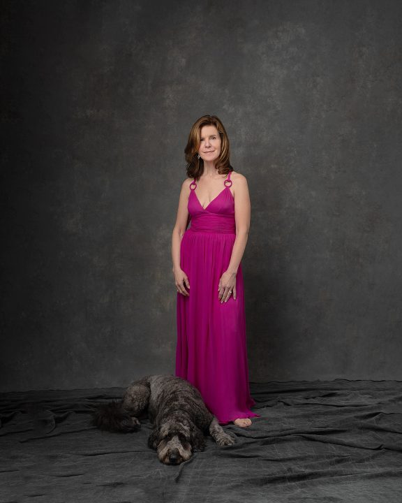 A portrait of Debra, wearing a long pink dress with her dog at her feet, during her photo session for The Over 50 Revolution
