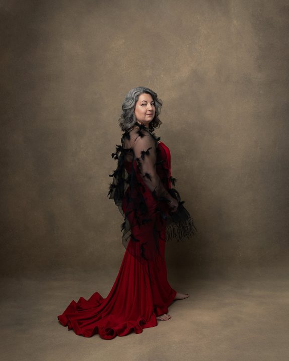 Portrait of Rebecca for The Over 50 Revolution portrait campaign. She is wearing a red dress and a black sheer scarf, and turned away from the camera, looking over her shoulder.