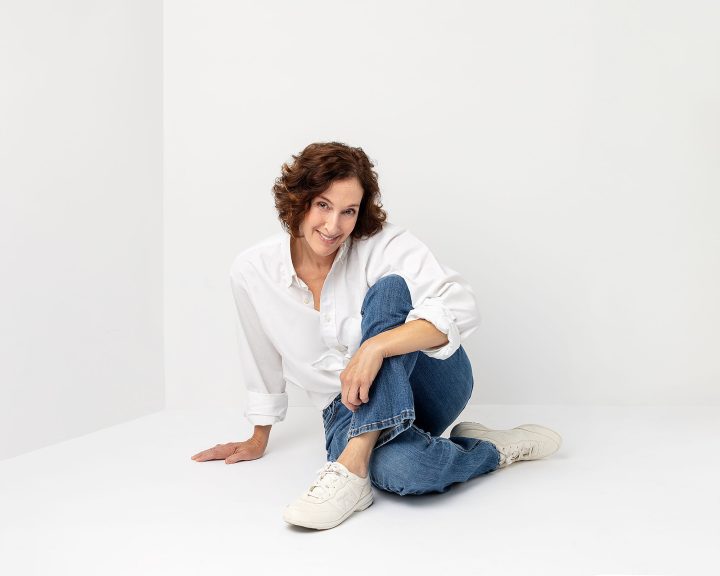 A portrait of Sandy, sitting on the floor, wearing a white shirt and jeans, from her session for The Over 50 Revolution