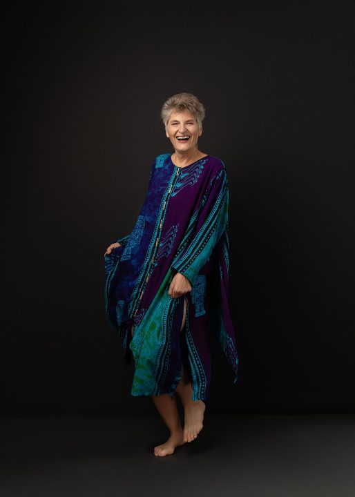 A portrait of Sue from her photo session for The Over 50 Revolution. She is wearing a colorful dress, dancing in front of a black background. 