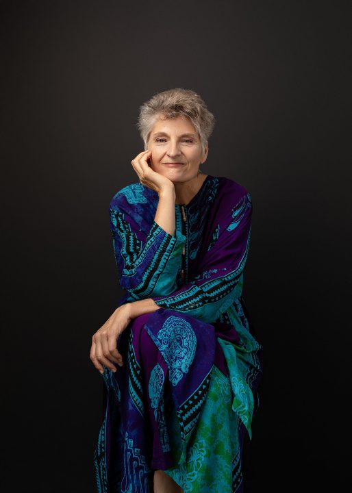 A portrait of Sue for The Over 50 Revolution. She is seated, wearing a colorful dress, in front of a black background.