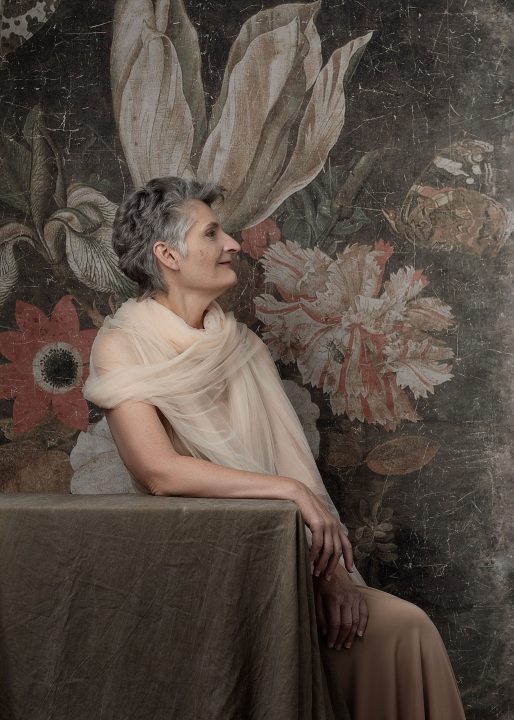 A profile portrait of Sue from her session for The Over 50 Revolution. She is wrapped in tulle in front of a floral backdrop.