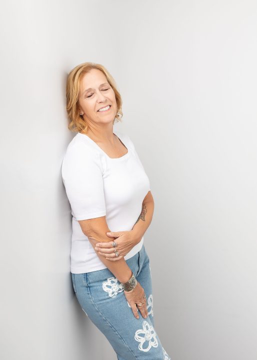 Portrait of Wendy for The Over 50 Revolution. She is wearing white, leaning against a white background, happy, with her eyes closed.