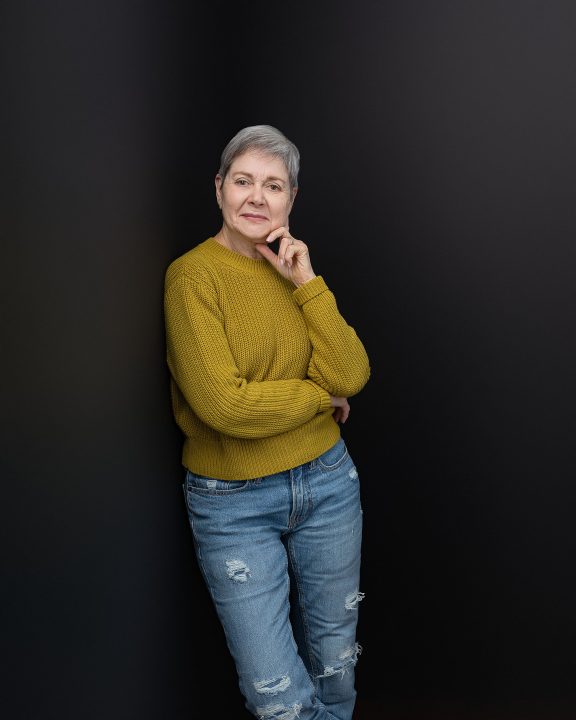 Portrait of artist Jill Fischman, leaning against a black wall, wearing a sweater and jeans