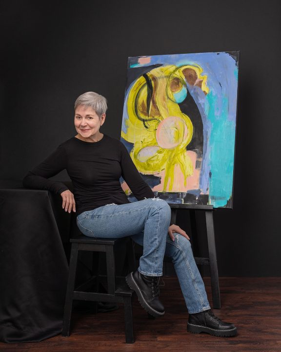 A portrait of artist Jill Fischman, seated, with her painting, in front of a black background