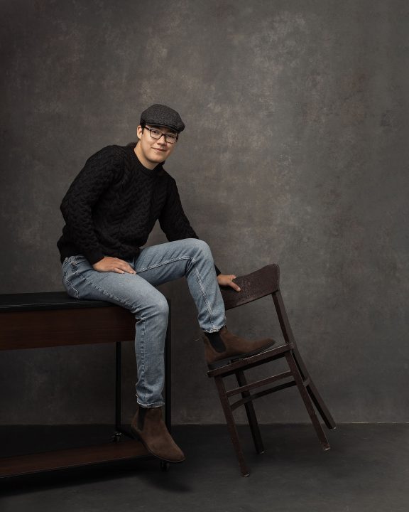 Portraits to celebrate 19 - David sitting on a desk, tipping a chair with his feet